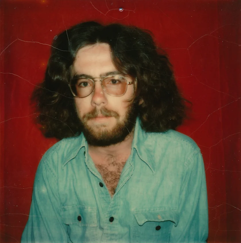 (Image: A colour photograph against a burgundy background of a young man, early twenties with shoulder-length wavy brown hair, thick beard and moustache, wearing tinted aviator lenses and a faded denim work shirt open at the neck.)