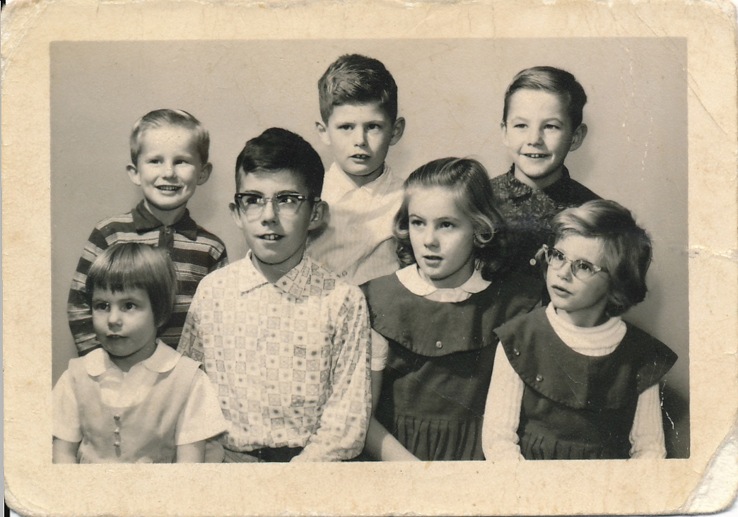(Family photo against a plain white background. From left to right on the bottom row is Shirley Ann, a young red-haired girl, Bruce aged 12 in glasses with binaural temple-mount hearing aids, Susan with long blond hair, Maureen with cat's eyes glasses and shorter hair. In the back row, a smiling Terry, serious Mike and grinning Allan.)