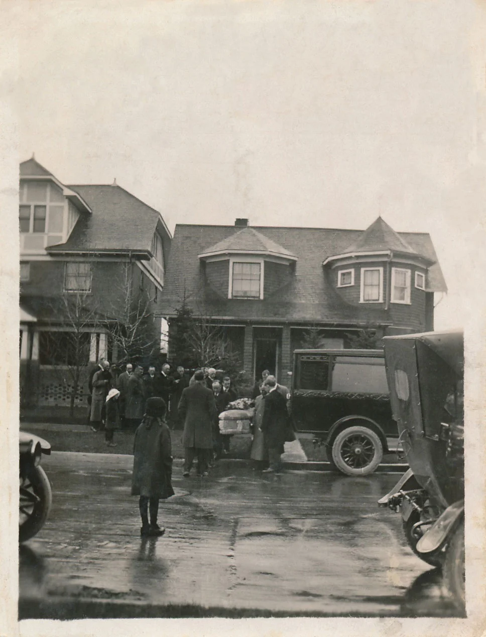 Family picture 1919 (Image: the funeral of Lavinia Begg as her casket is taken by pallbearers from the family home at 815 - 14th Ave. S.W. to a waiting hearse. Family members visible in the background include Robert Begg, Lavinia's Sr.'s sister Martha and her husband Hiram Worden. In the foreground, young Lavina Jr. in a long dark skirt standing in the rain-slicked street facing the hearse says goodbye to her mother. Printed on the back of the photograph is a single word, "Mother".)