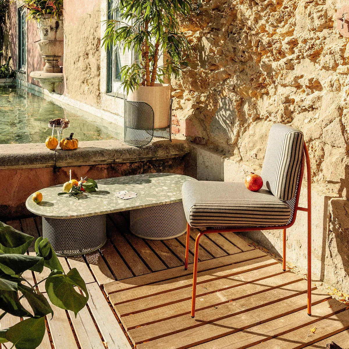 An outdoor lifestyle scene featuring a striped Riviera chair, a green terrazzo coffee table with a perforated metal base, and lush greenery by a stone pool.
