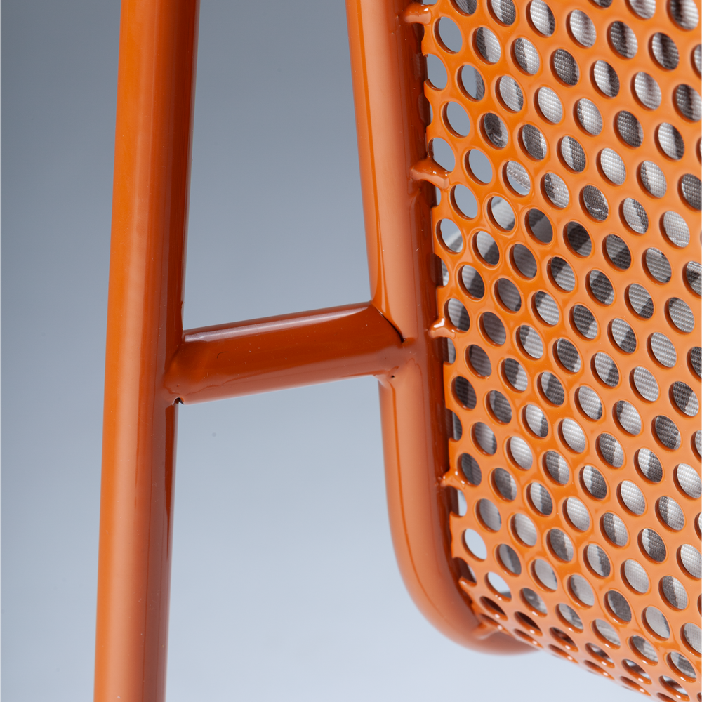 A macro close-up of a terra cotta orange powder-coated metal frame meeting a perforated mesh backrest on a contemporary chair.