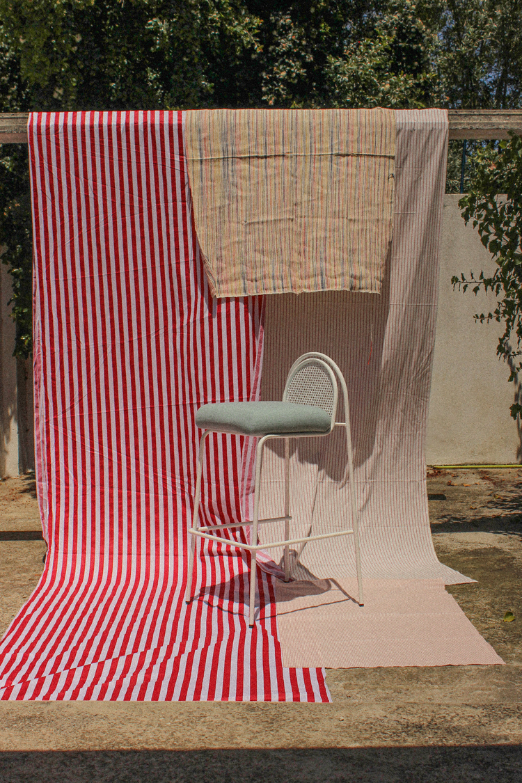 A vertical outdoor shot of a white metal bar stool with a solid sage green cushion, placed against vibrant red and white striped hanging textiles.