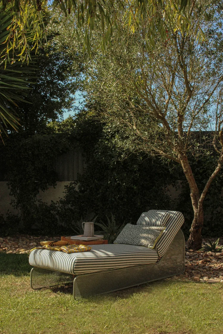 Lifestyle shot; single daybed in a garden setting; striped upholstery; textured accent pillow; shaded by olive trees; natural stone ground.