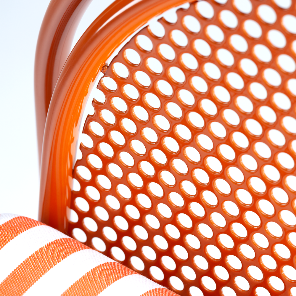 A macro shot showing the junction where the perforated metal backrest meets the orange frame, with a partial view of the striped fabric cushion in the lower corner.