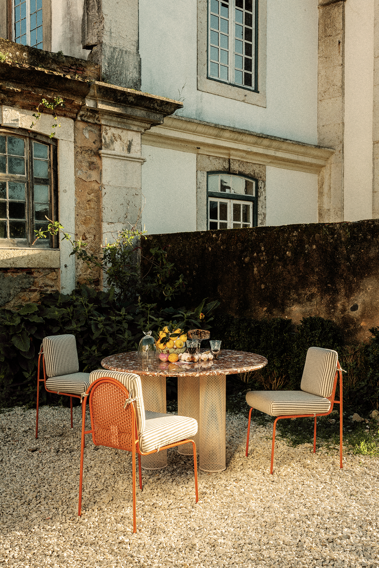 Three upholstered chairs with orange frames flanking a circular white terrazzo dining table supported by three perforated cream metal pillars.
