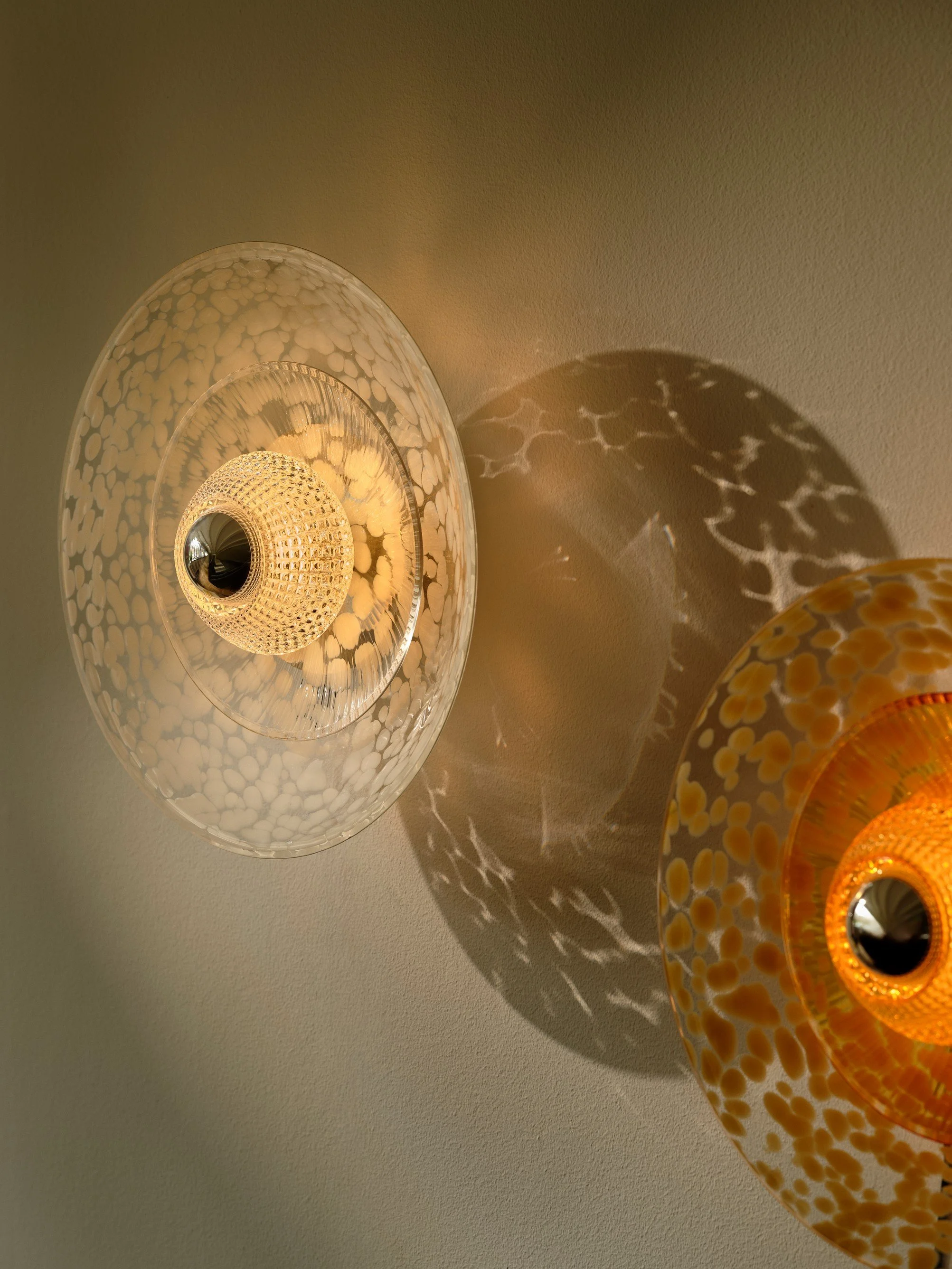 Clear and Amber Disco Rama sconces side by side, casting overlapping dappled light across a plaster wall.