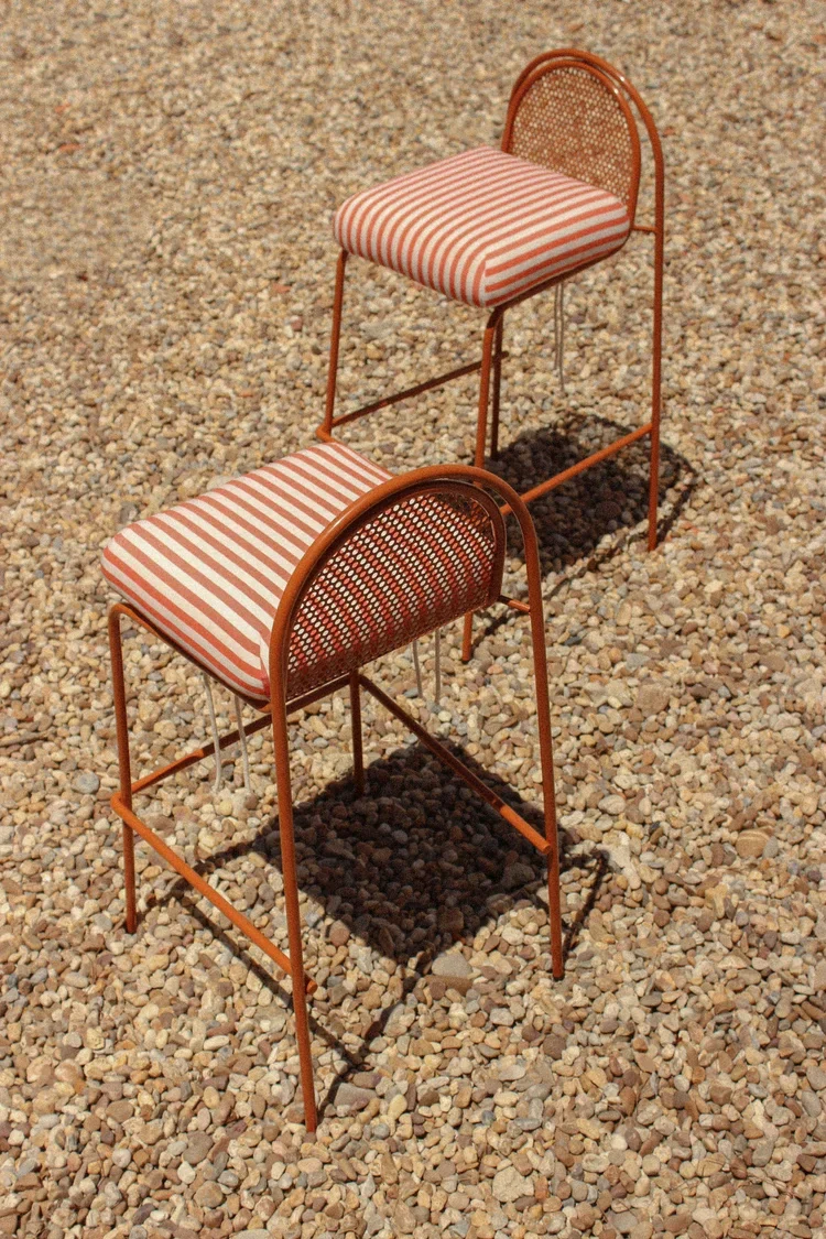 A high-angle shot of two orange metal bar chairs with striped cushions, placed on a gravel ground, emphasizing the geometric shape of the frames and shadows.