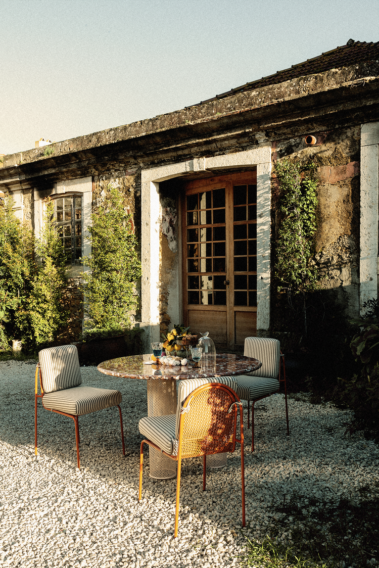 A luxury outdoor dining set featuring three striped Riviera chairs and a circular red marble-top table with a perforated pedestal base, set in a gravel Mediterranean courtyard.