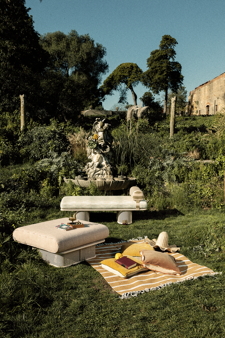 A lifestyle photograph of a minimalist orange metal bench with a striped cushion, placed on a sun-dappled concrete patio next to a white wall and green plants.