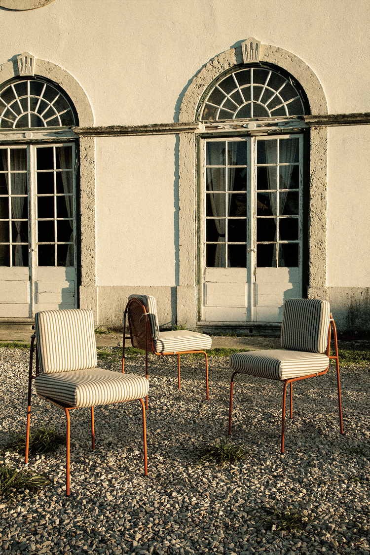 Three Riviera dining chairs with orange frames and striped upholstery arranged on a gravel courtyard in front of a white stone building with arched French doors.