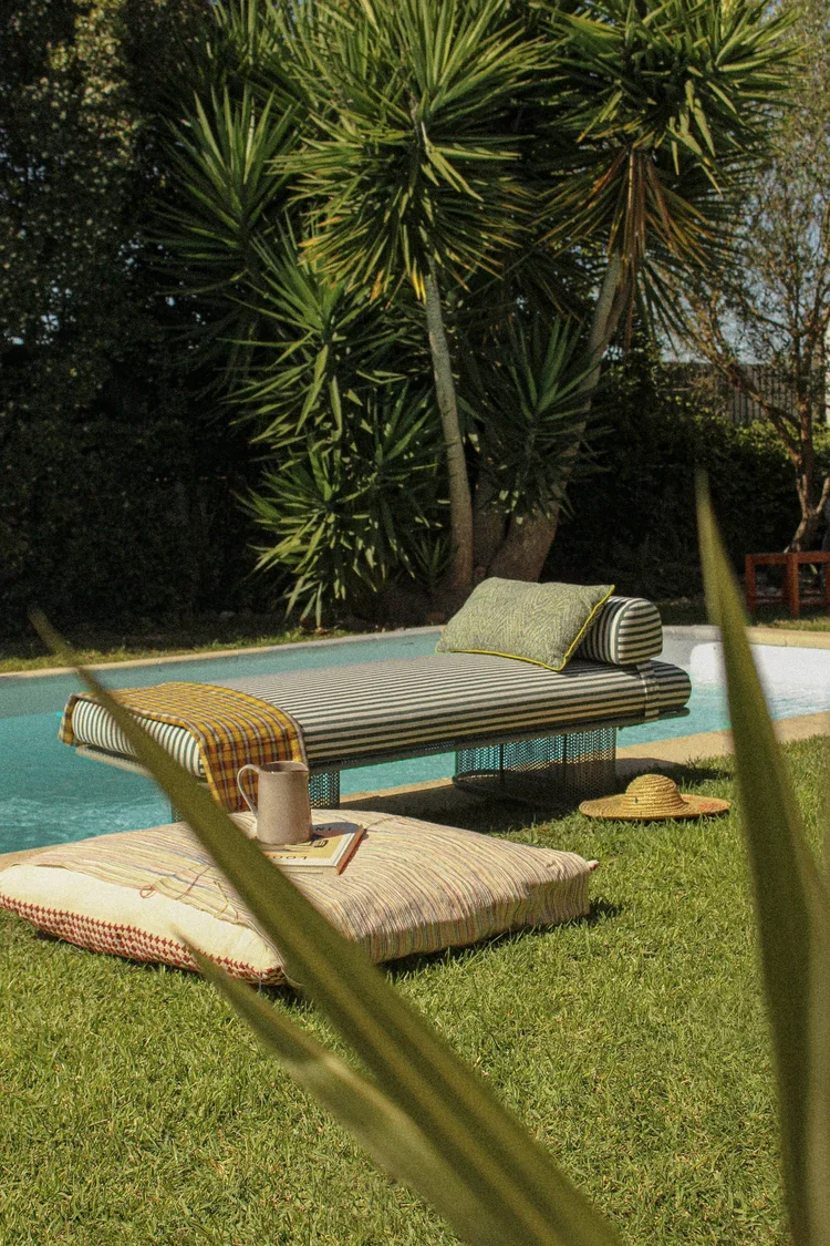 Riviera outdoor bench with striped cushion placed by a swimming pool surrounded by greenery
