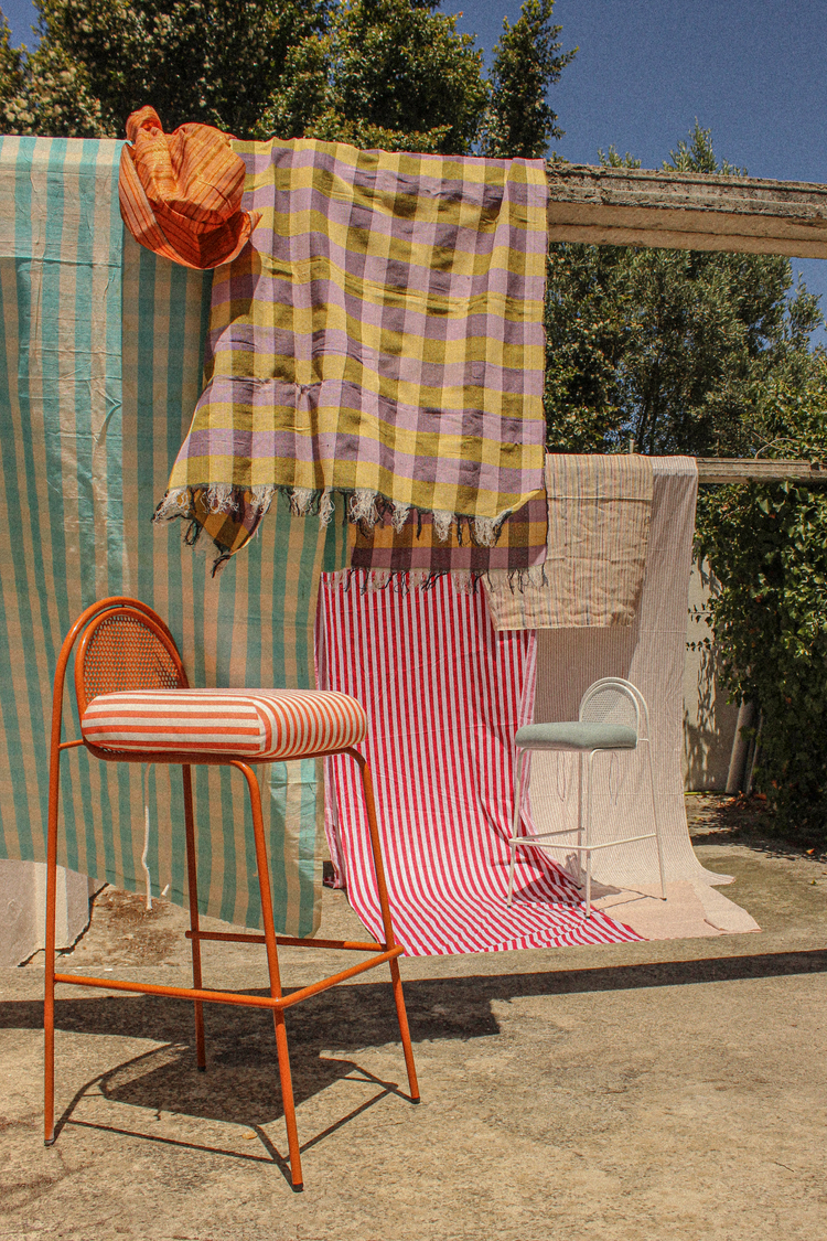 A wide outdoor shot featuring various patterned textiles (checkered, striped) hanging from a beam, with two bar chairs in the foreground on a concrete patio.