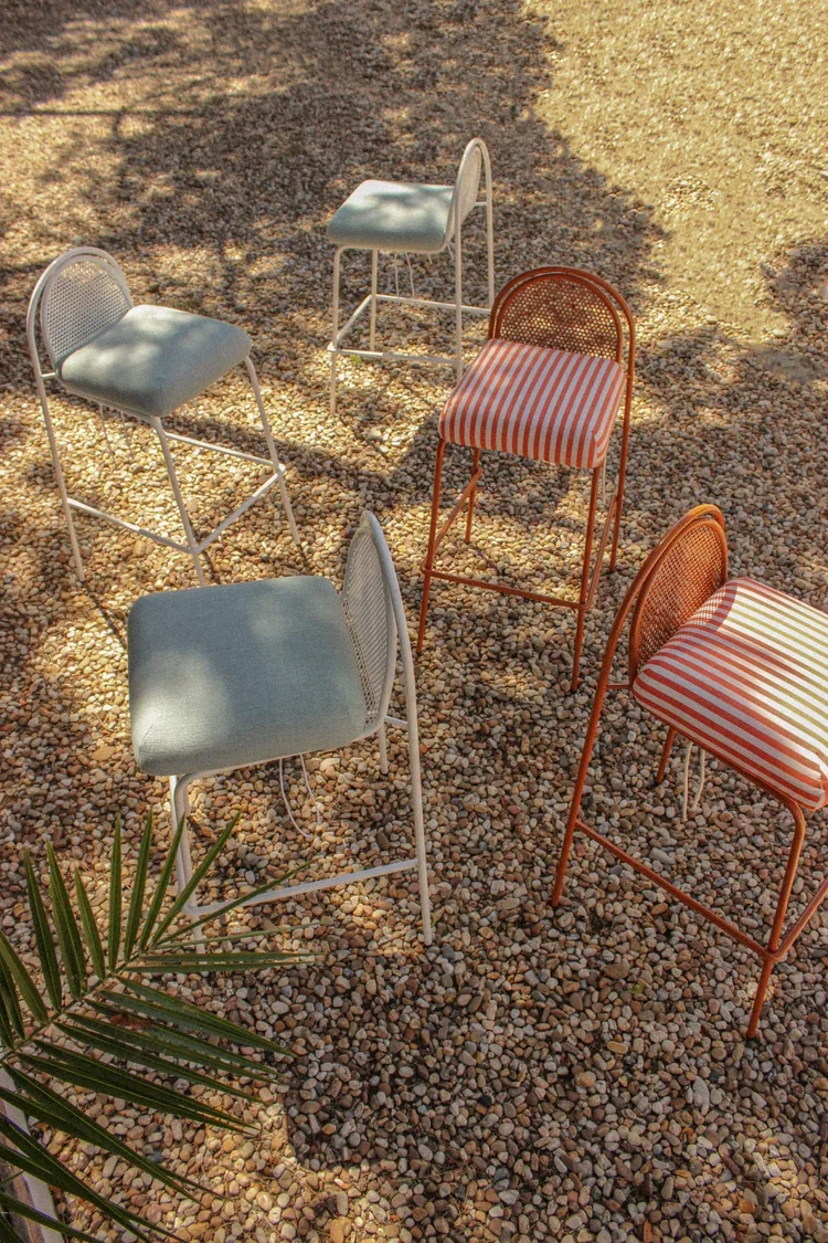 An elevated lifestyle shot of five bar stools in orange and white finishes, scattered across a light-colored gravel surface with soft afternoon shadows.
