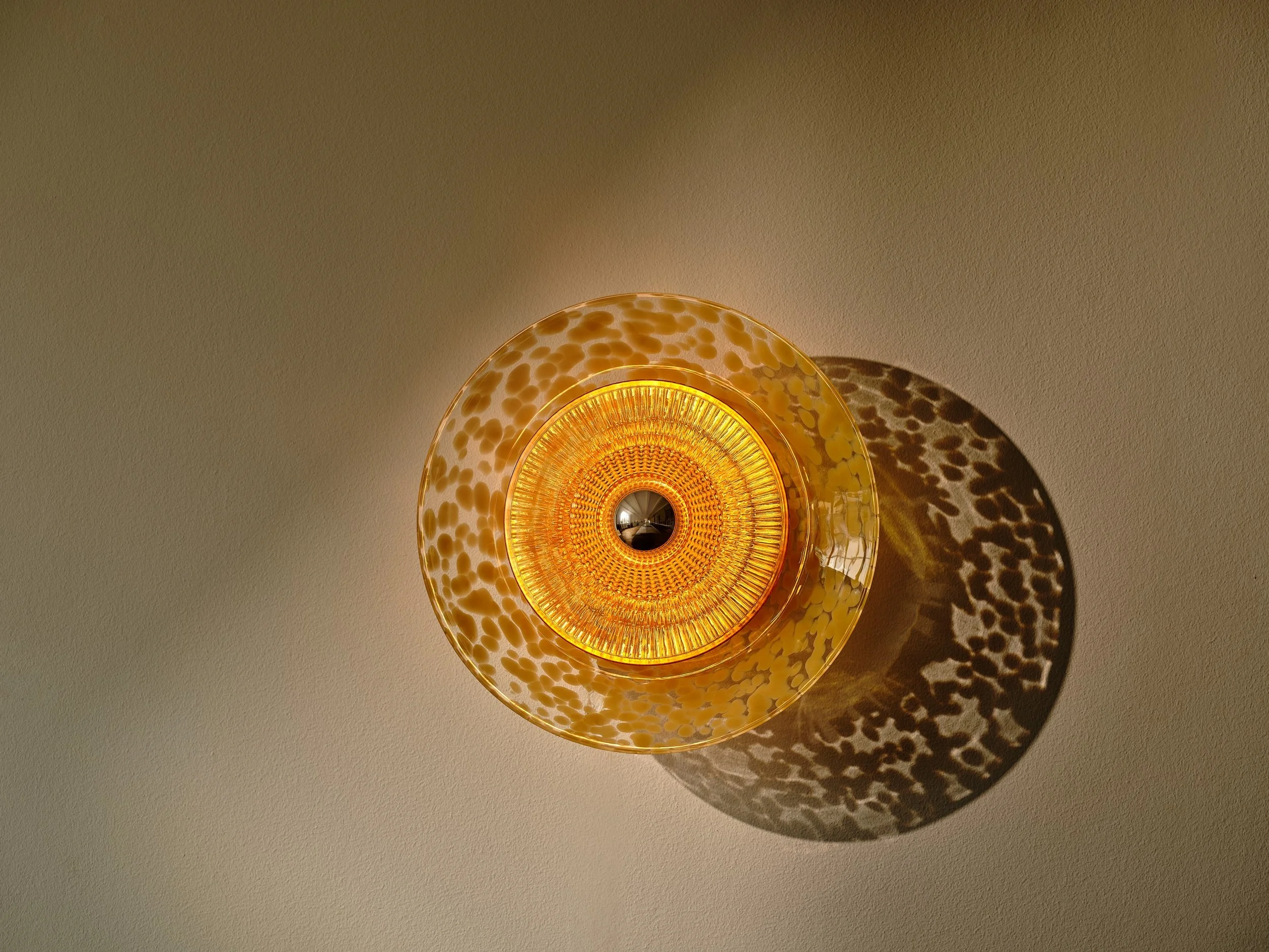 Amber Disco Rama sconce illuminated on a warm plaster wall, projecting a golden dappled shadow halo.