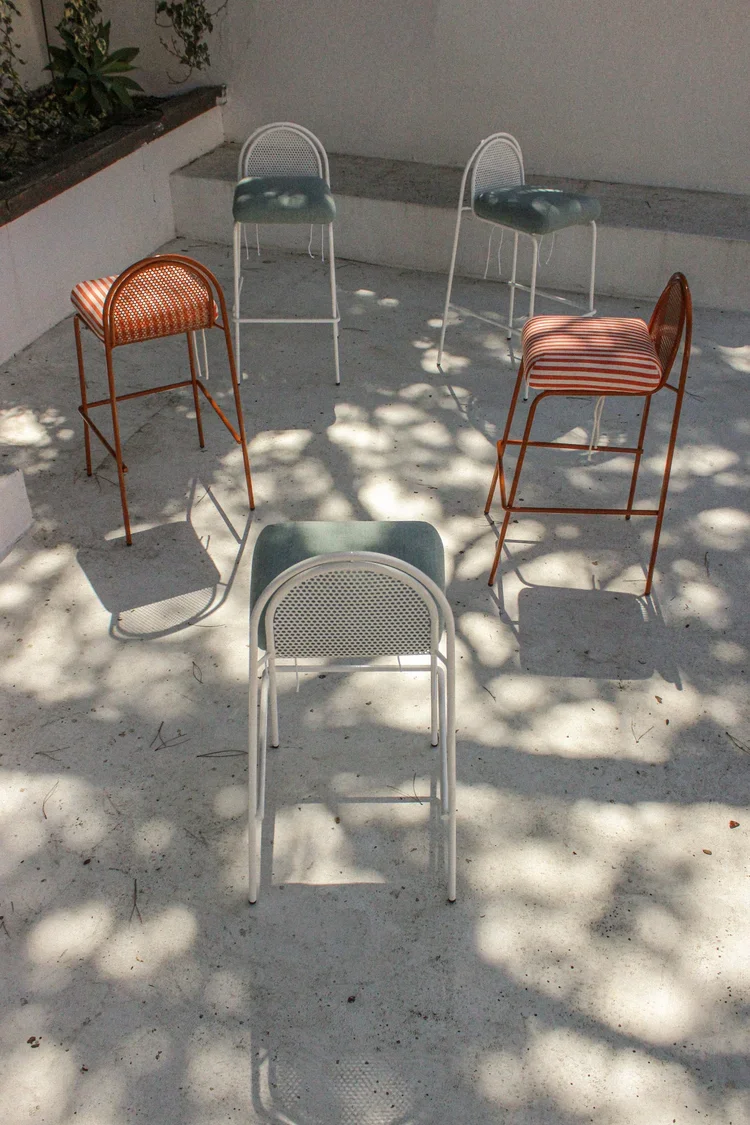 A lifestyle photograph of four bar chairs (two orange with striped cushions and two white with sage green cushions) arranged on a sun-dappled concrete terrace.