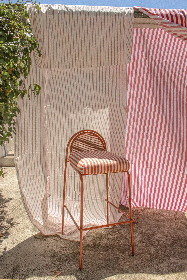 A vertical outdoor shot of a burnt-orange metal bar chair with a red and white striped seat cushion, positioned against a background of striped fabric backdrops.
