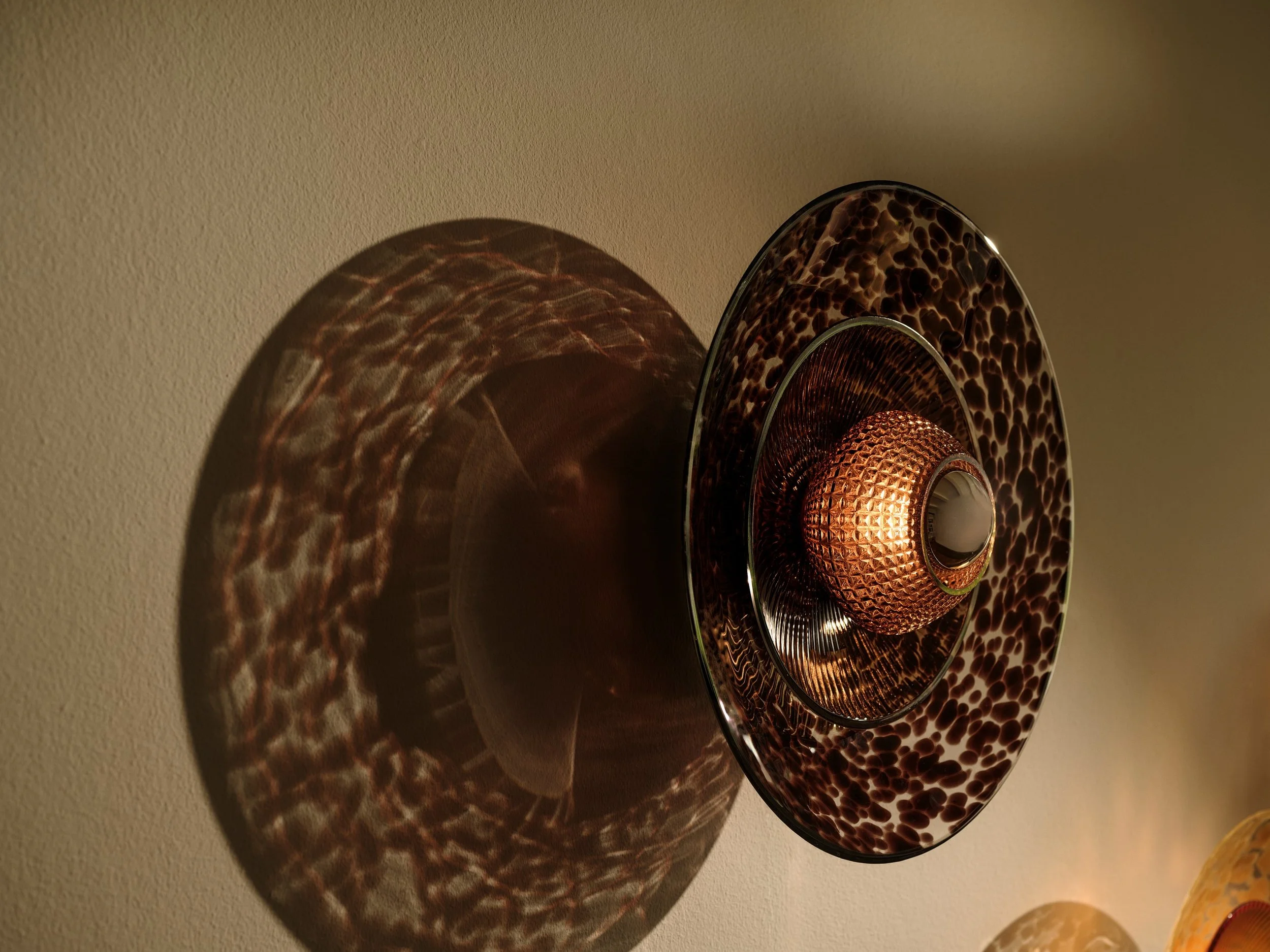 Brown Disco Rama sconce lit on a plaster wall, casting a velvety tortoiseshell shadow pattern.