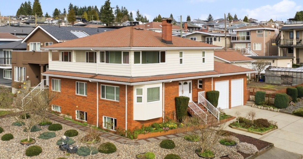 A large modern house with multiple gabled roofs, white siding, and stone accents, surrounded by tall trees in a forest setting. The house has a well-lit exterior, a driveway, a basketball hoop, a backyard patio with outdoor seating, and landscaped plants.