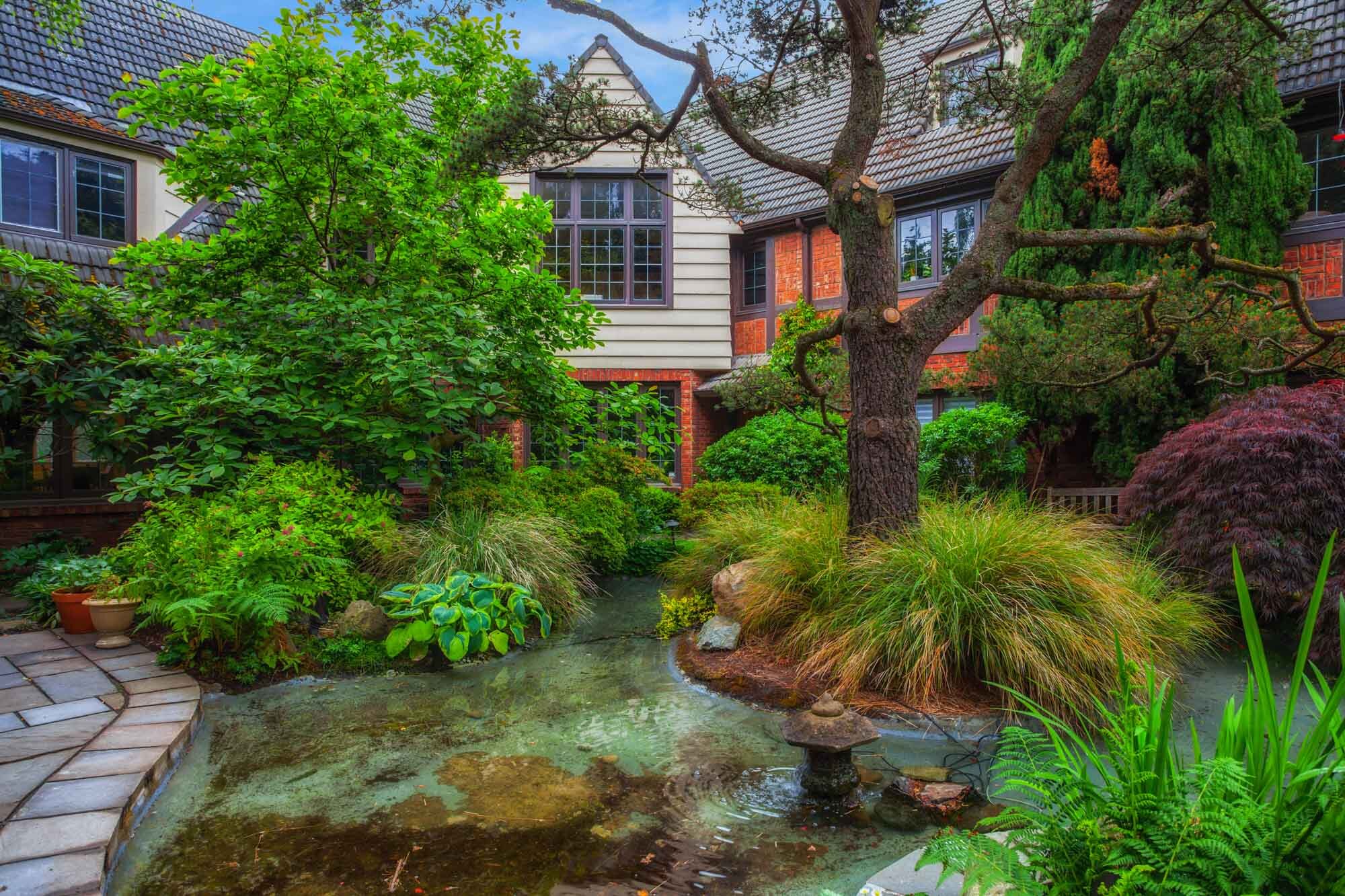  image description: exterior of storybook tudor with garden, trees, pond, and walkway 