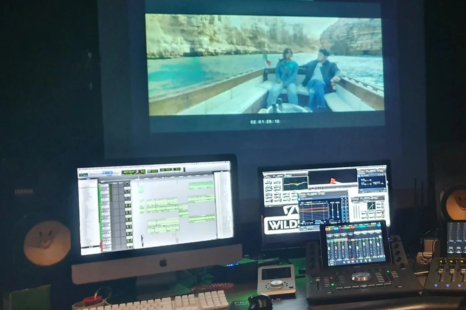 A music production studio with multiple monitors displaying audio editing software, a DJ controller, a keyboard, speakers, and a tablet.