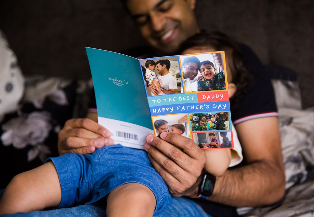 In-Home Family Photo Session In Wimbledon with boy holding a fathers day greeting card while sitting on dad's lap