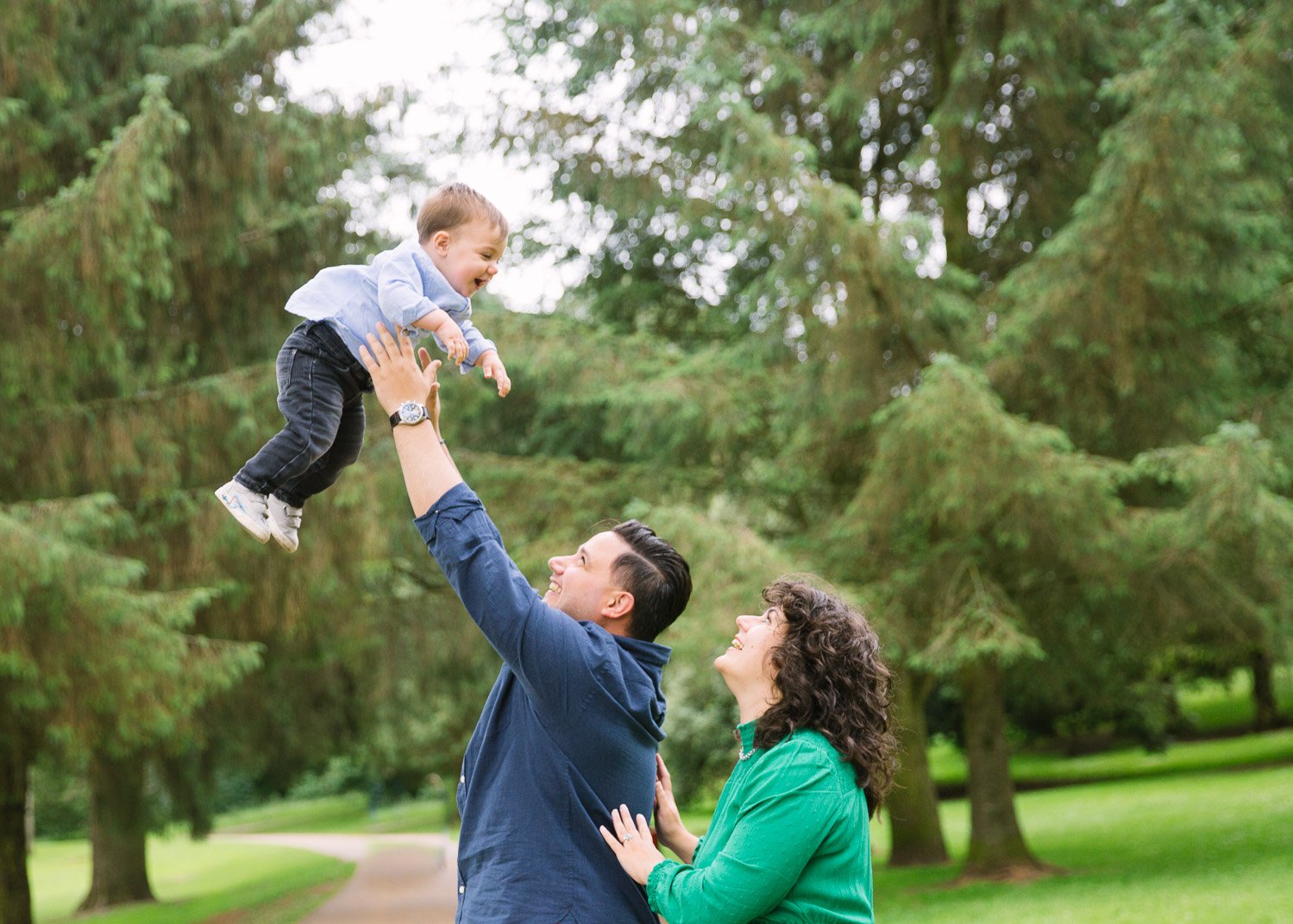 One year old laughing while flung in air by dad and mum looks on lovingly in outdoor family photoshoot in wimbledon