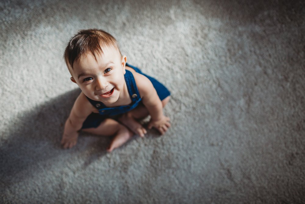 Wimbledon Family Photographer captures a one year old child sitting on the carpet looking up and smiling