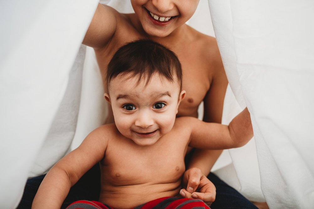 Wimbledon Family Photographer captures siblings laughing under a blanket in a home photoshoot