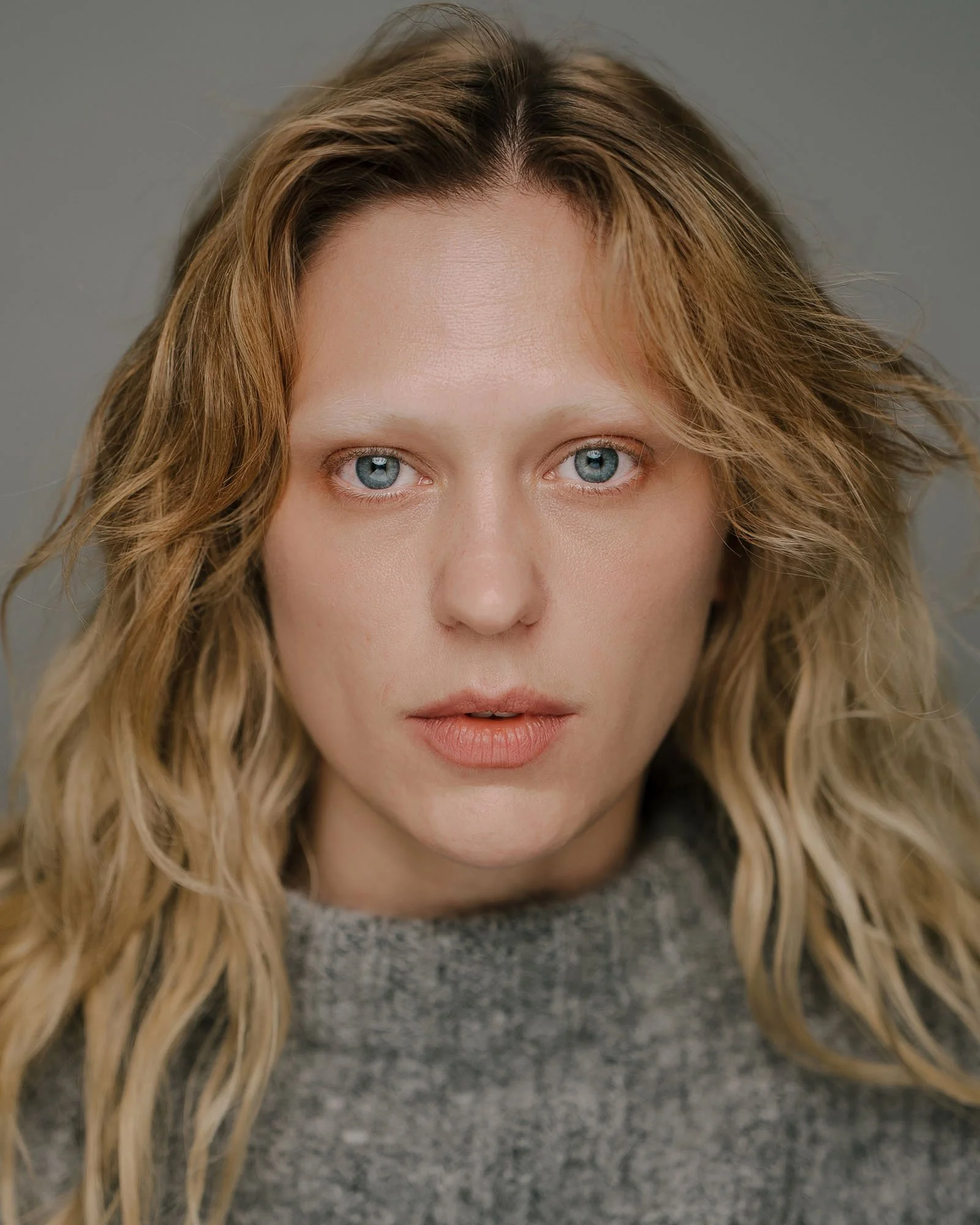 Actor headshot of a woman with long blond curly hair in a grey knitted jumper against a textured grey backdrop.