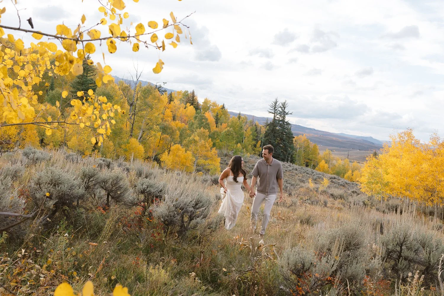 A man and woman hold hands and frolic through a sagebrush meadow surrounded by yellow aspens in the fall in Colorado mountains, engagement photographer.