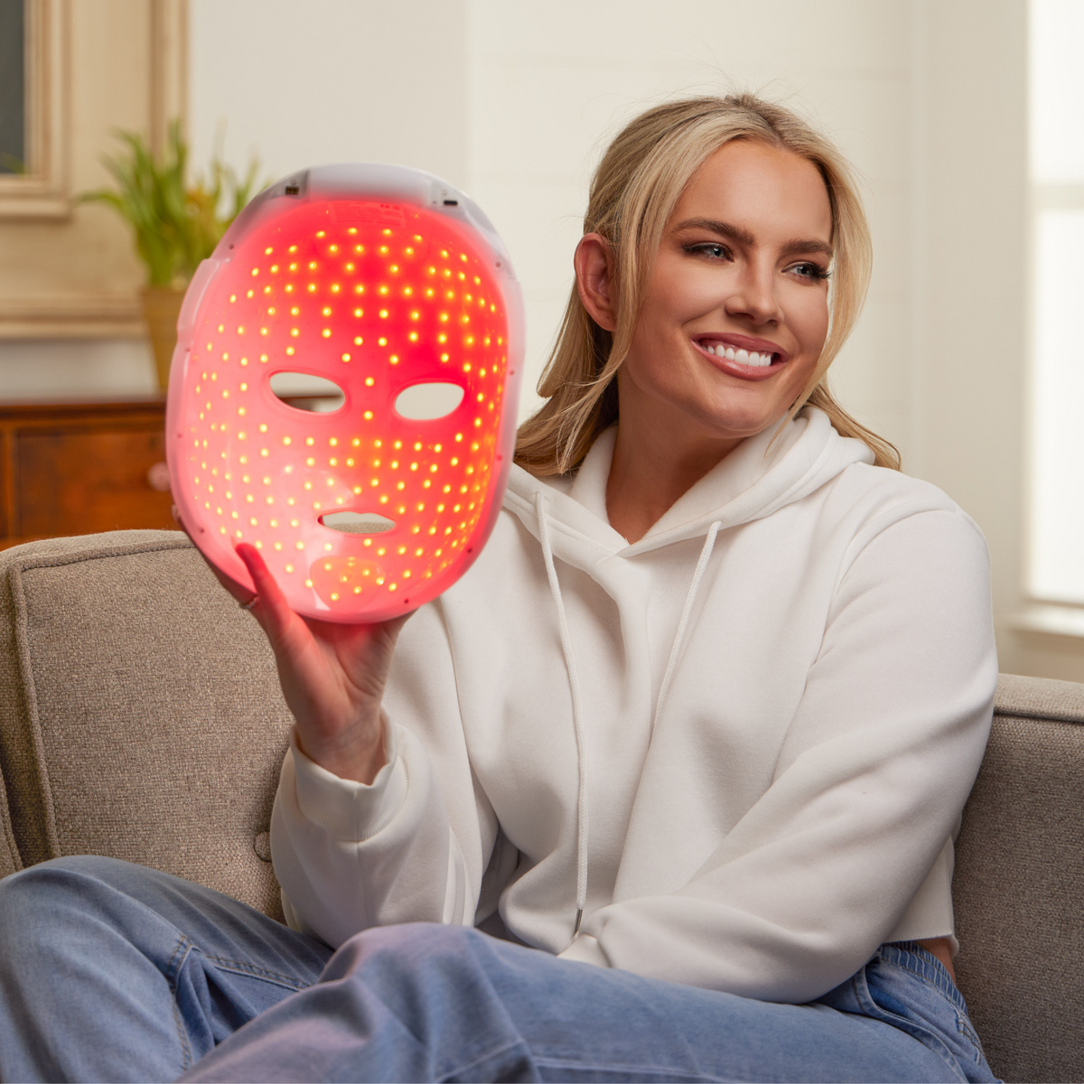 Woman sitting on a sofa at home holding a red-lit LED face mask during an evening spa routine