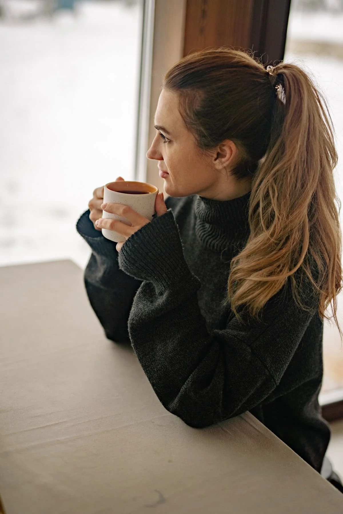 Woman in a heavy sweater holding a mug beside an apartment window on a cold day