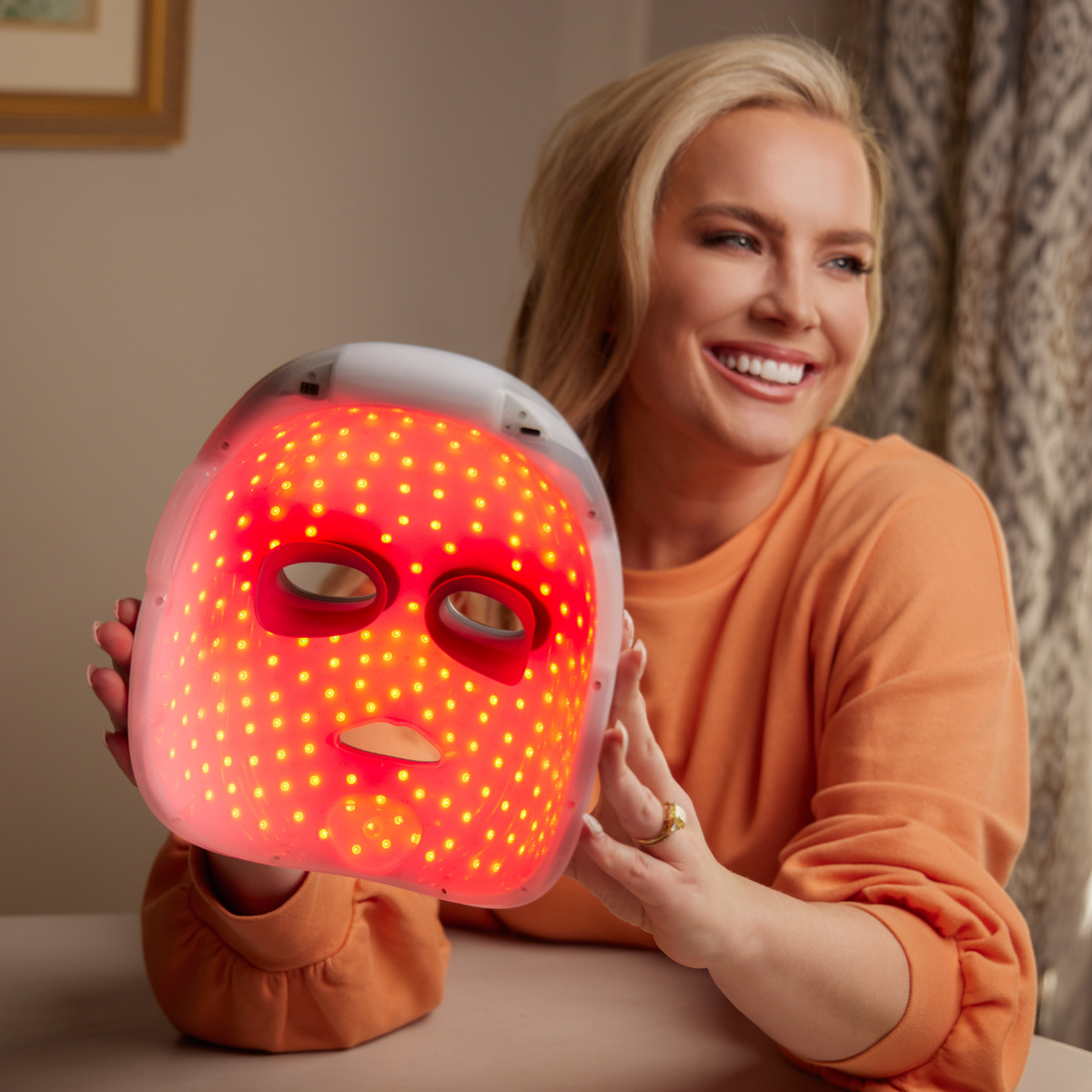 Close view of an LED face mask glowing red, held up in a warmly lit living room