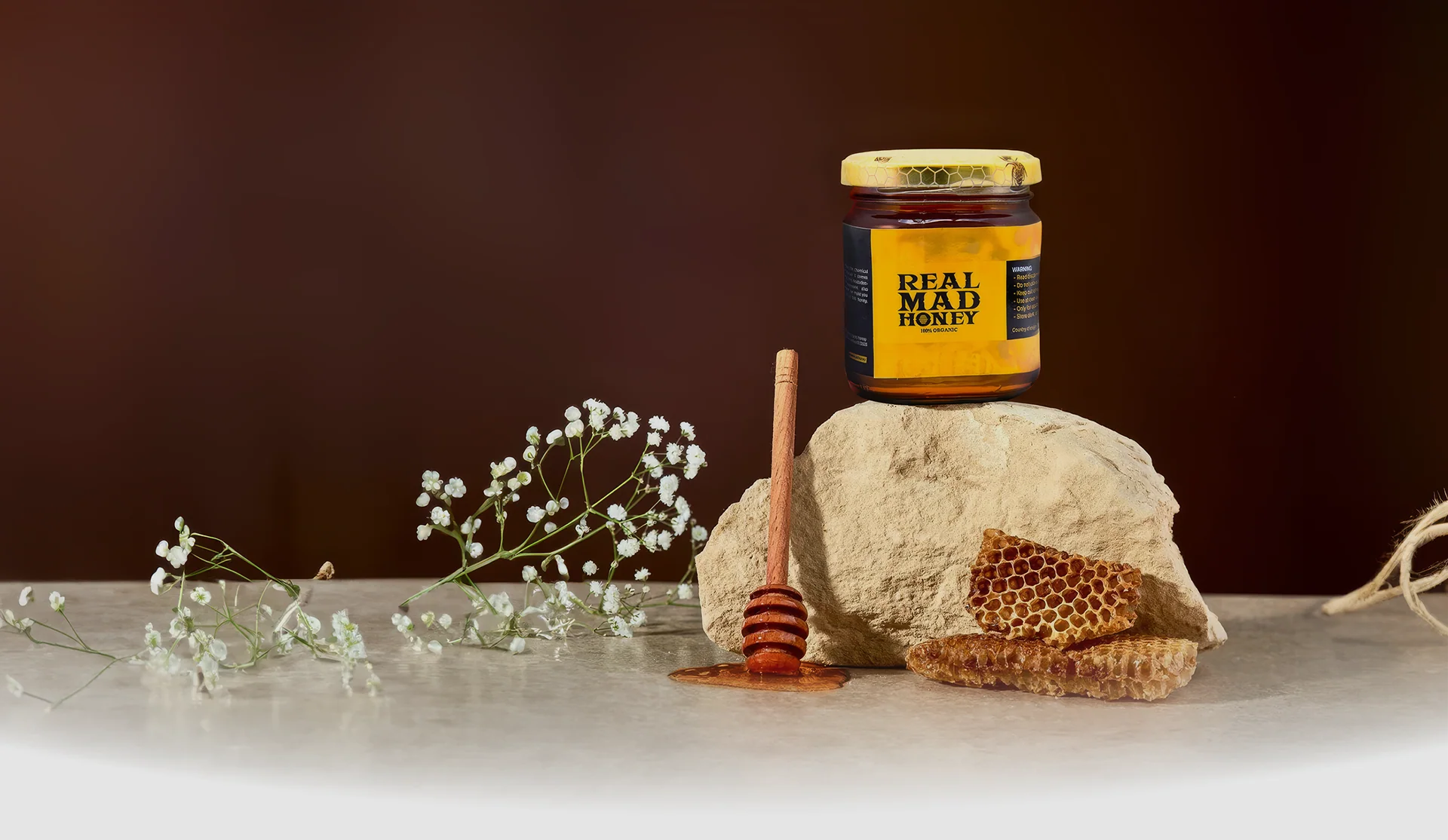 Real Mad Honey jar displayed on pale stone beside honeycomb, a wooden dipper and small white flowers