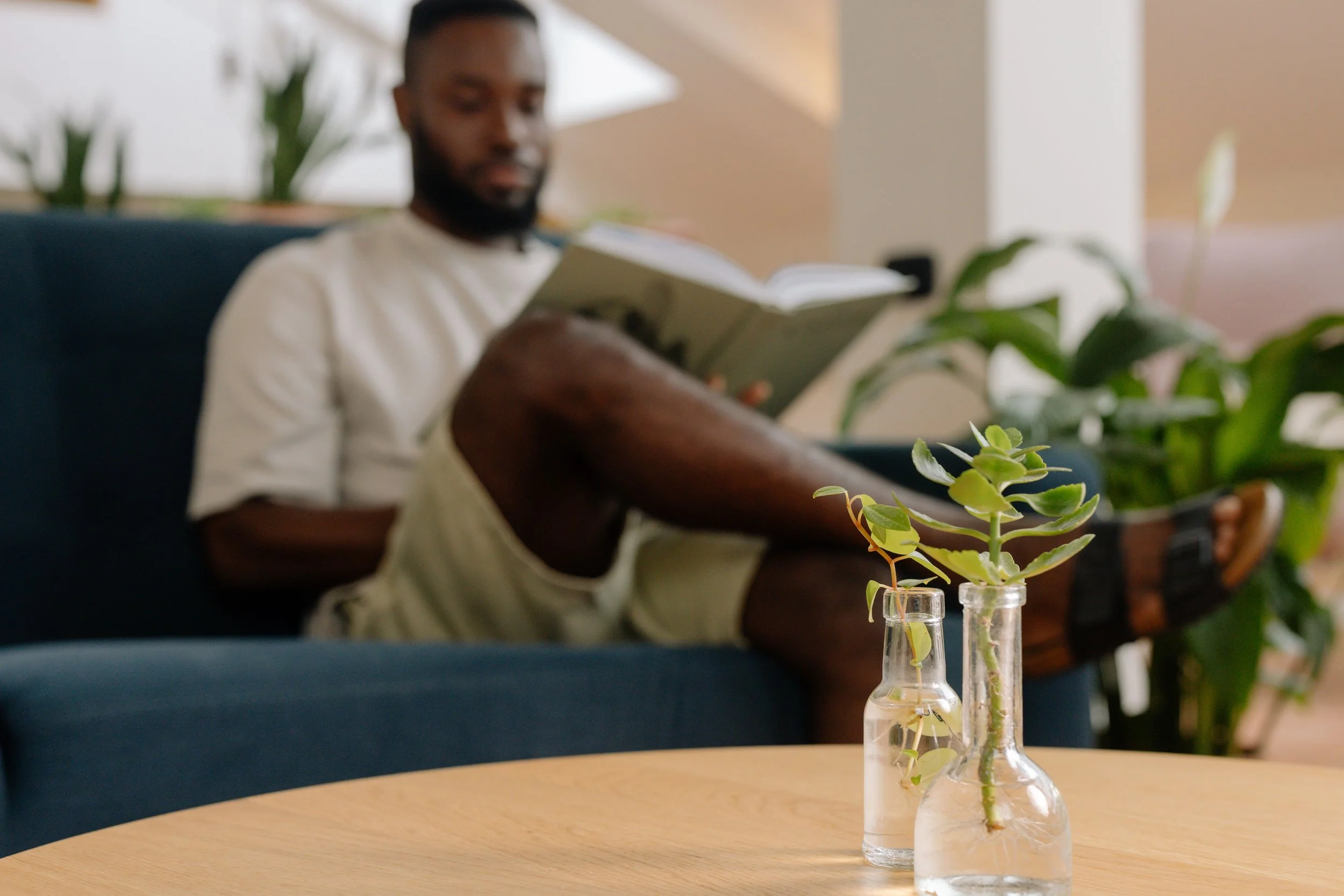 Black man relaxing on a blue sofa while reading a book in a bright, plant-filled room.