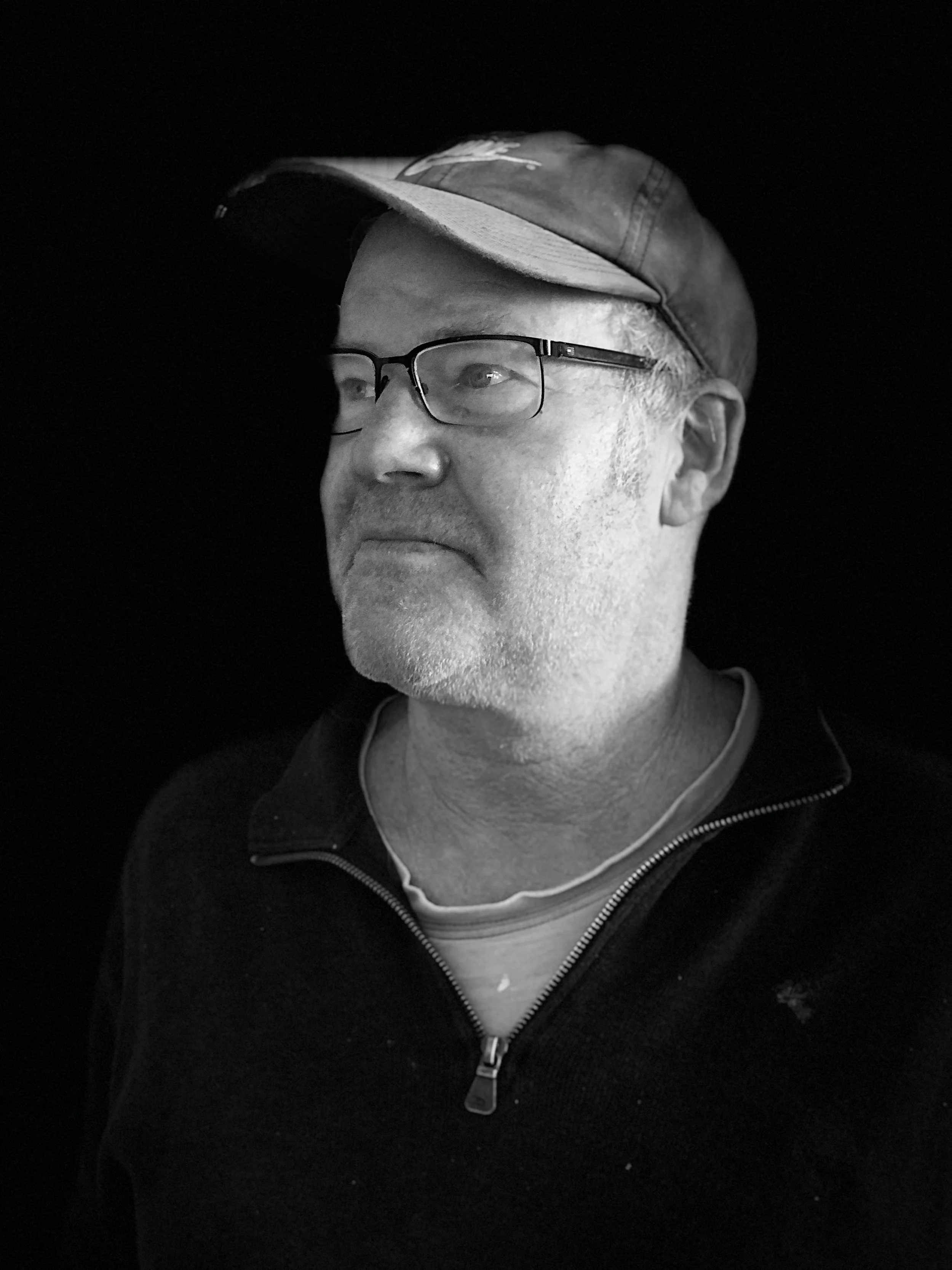 Black and white photo of Tim Barlow. Tim wears a cap and glasses and has grey stubble around his mouth, chin and neck