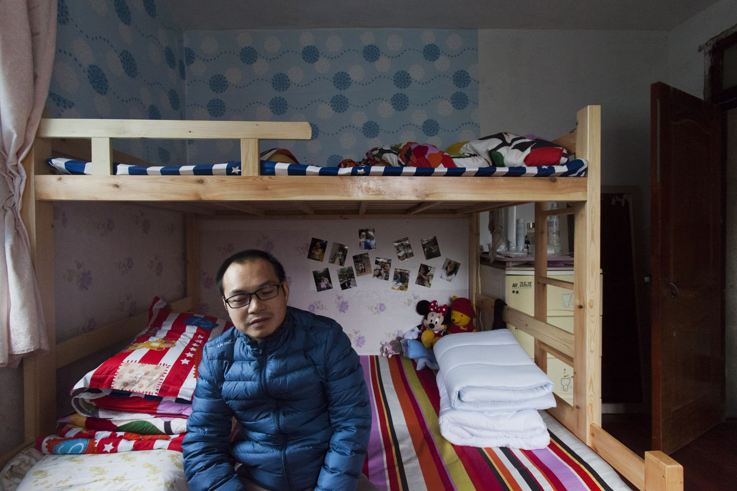 A father sits on the family bunk bed in their one room apartment. Photos of his family are taped to the wall behind him. Guangfu Road. Shanghai, China. 2015.