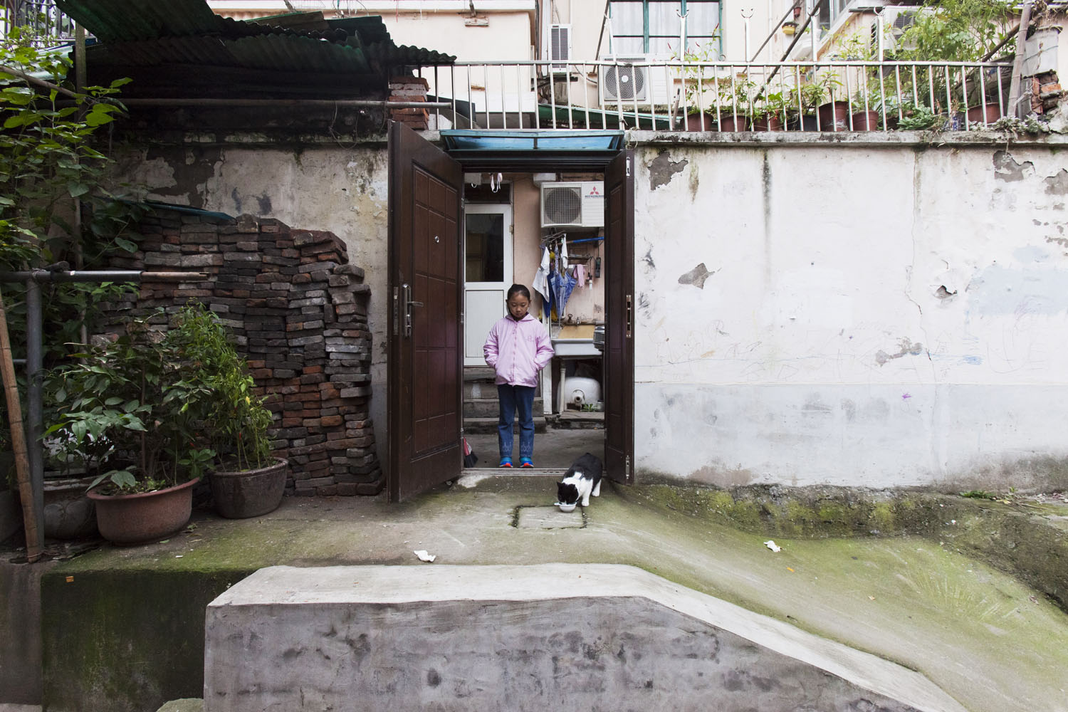  Young girl feeds a cat in front of her family's place of residence. Guangfu Road. Shanghai, China. 2015.