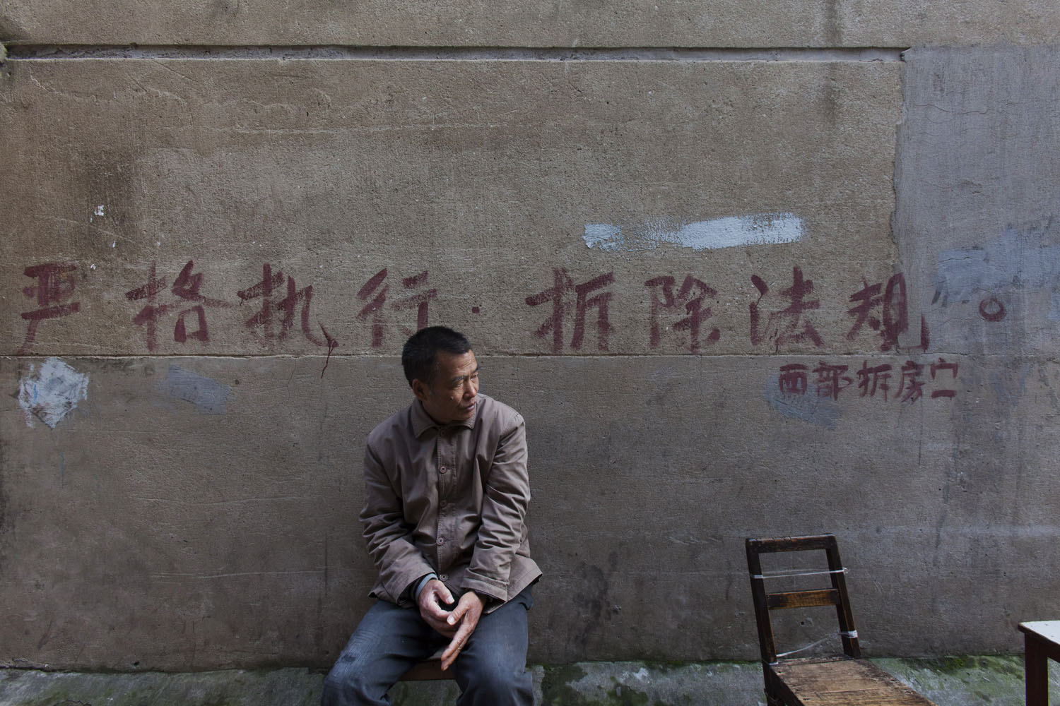 A man sits in front of a painted neighborhood notice that reads, "Abide by Housing Removal/Relocation Policy". Guangfu Road. Shanghai, China. 2015.