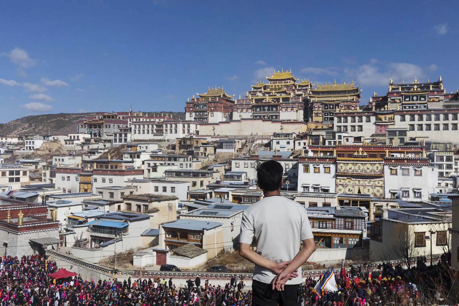 A man observes the crowd below him, gathered for the parade as it passes the front of JingXiang Village, at the base of the SongZangLin Monastary. Yunnan, China. March 2, 2018.