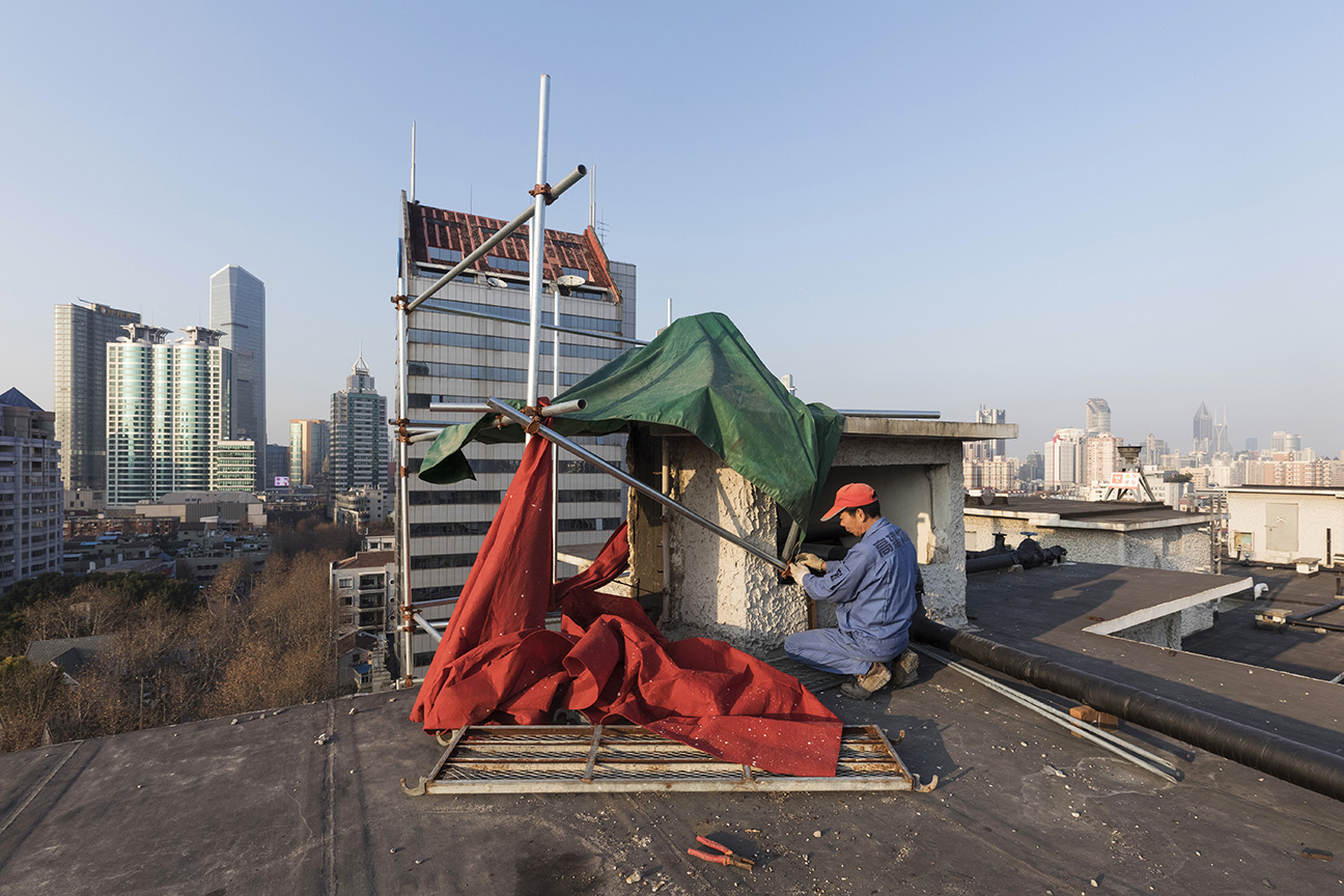 Worker takes down a tarp after finishing his day on a rooftop in Shanghai, China. 2018.