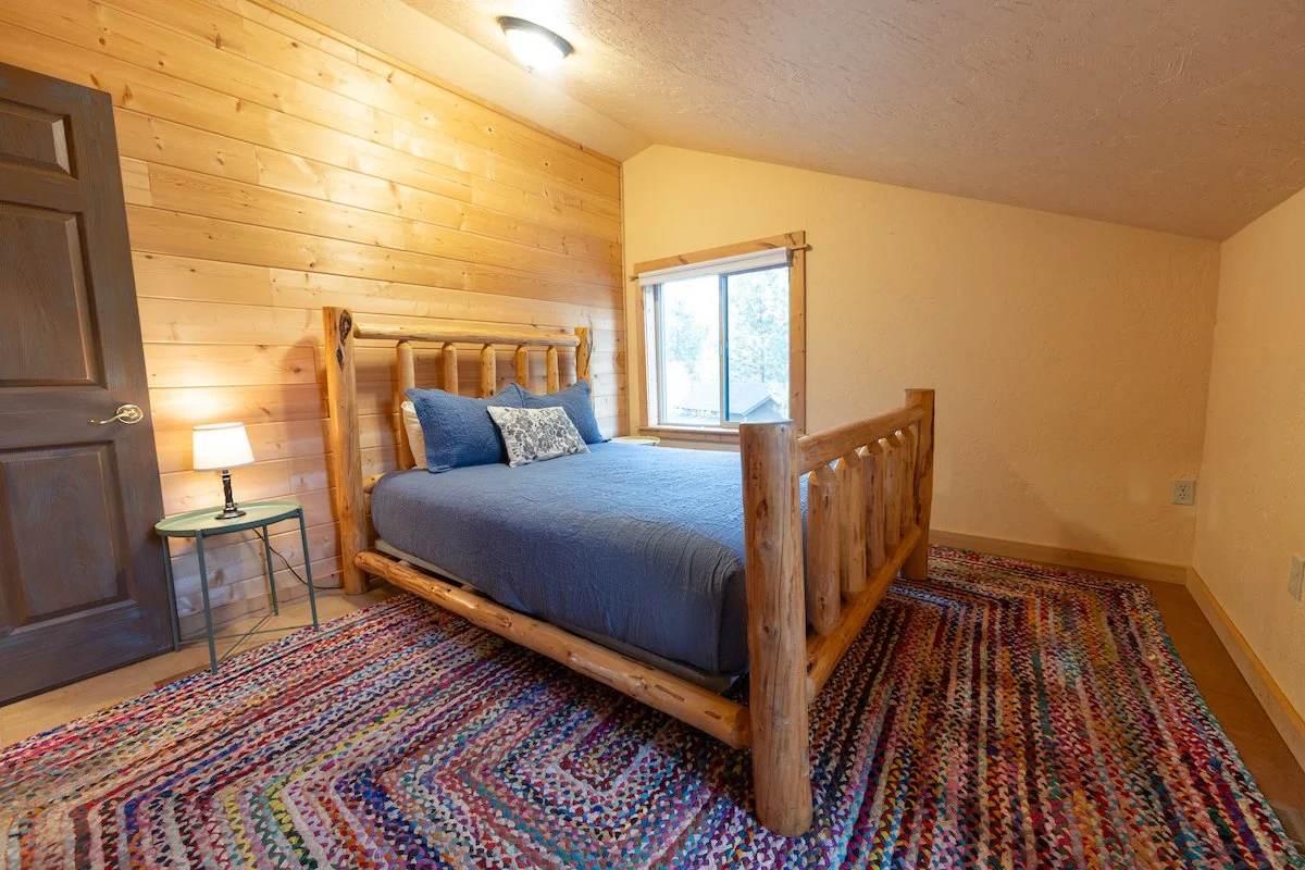 Bedroom with wooden walls, a wooden bed with blue bedding, a colorful braided rug, and a window with natural light.