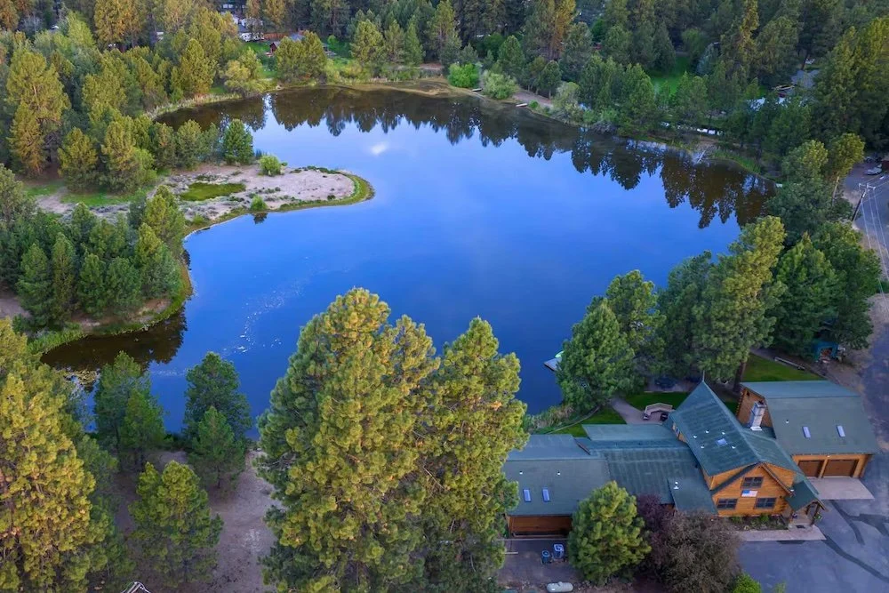 A scenic aerial view of a lake surrounded by dense trees and a cabin at the bottom right corner.
