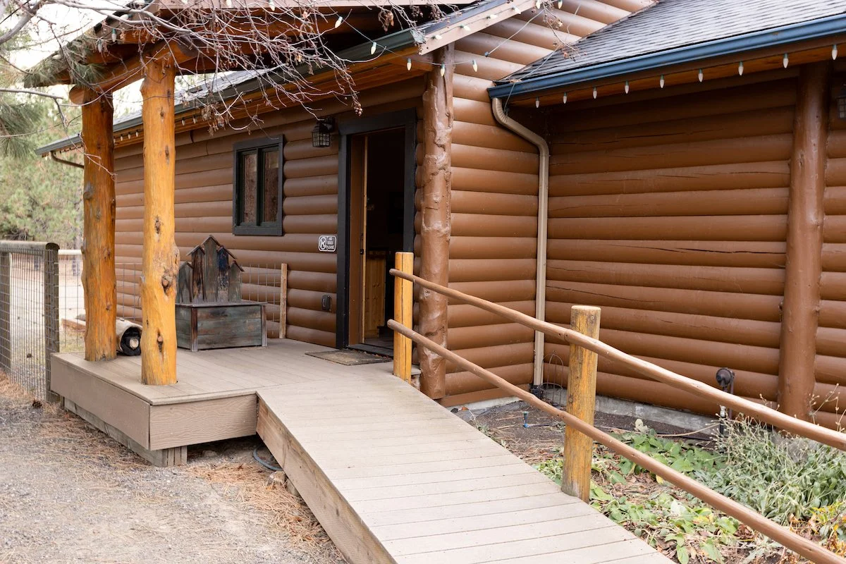 Front entrance of a log cabin with a small ramp, wooden railing, and string lights hanging from the roof, surrounded by trees.