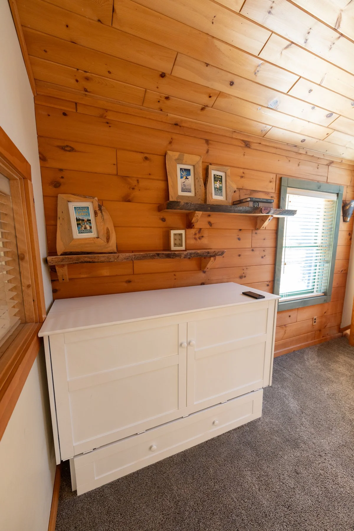 Room with wood-paneled sloped ceiling, white cabinet against wall, and two rustic wooden shelves holding framed pictures and books.