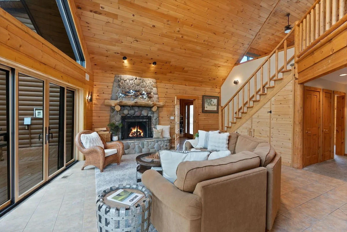 Living room with high wooden vaulted ceiling, stone fireplace, beige couches with pillows, wicker chair, sliding glass door, and staircase leading upstairs.