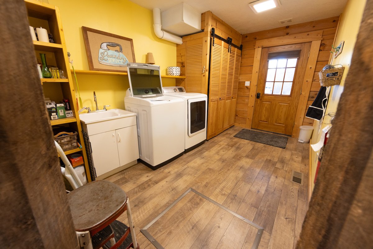 Laundry room with wooden walls and flooring, featuring a washer and dryer, a small sink, wooden cabinets, a door with a window, and shelves with laundry supplies.