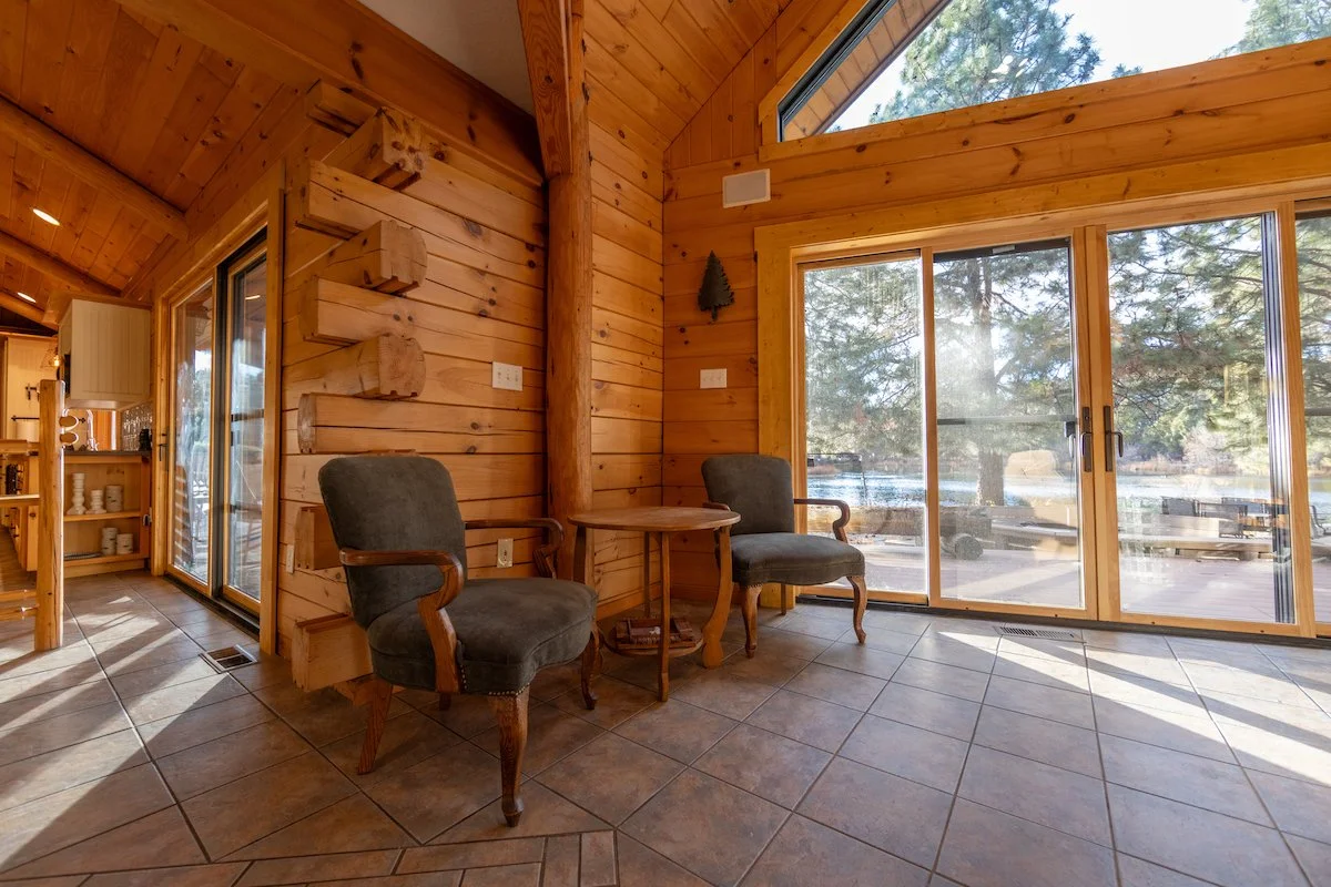 A cozy interior with wooden paneled walls, a large glass sliding door, and two upholstered armchairs around a small round table, overlooking a scenic outdoor view of trees and water.