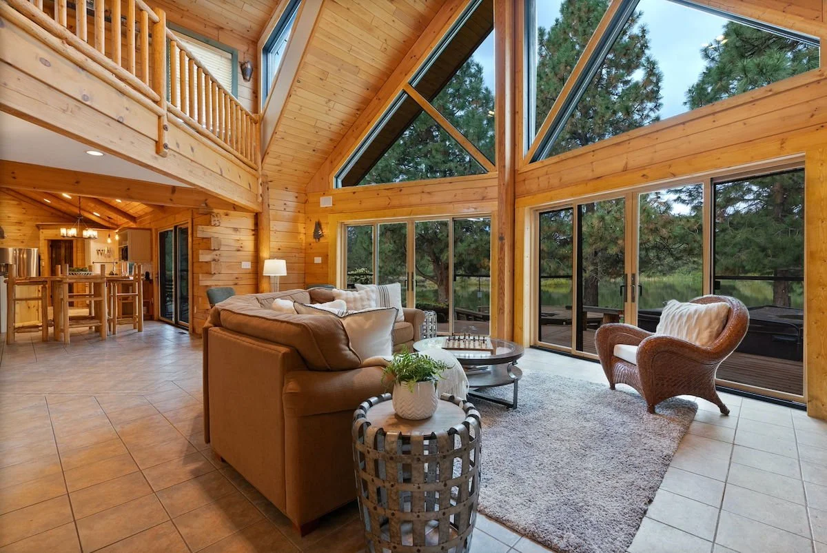 A spacious living room with wooden walls and large triangular windows showing trees outside. There's a beige sofa with pillows, a rattan armchair, a rug, and a coffee table. Sliding glass doors lead to an outdoor deck.