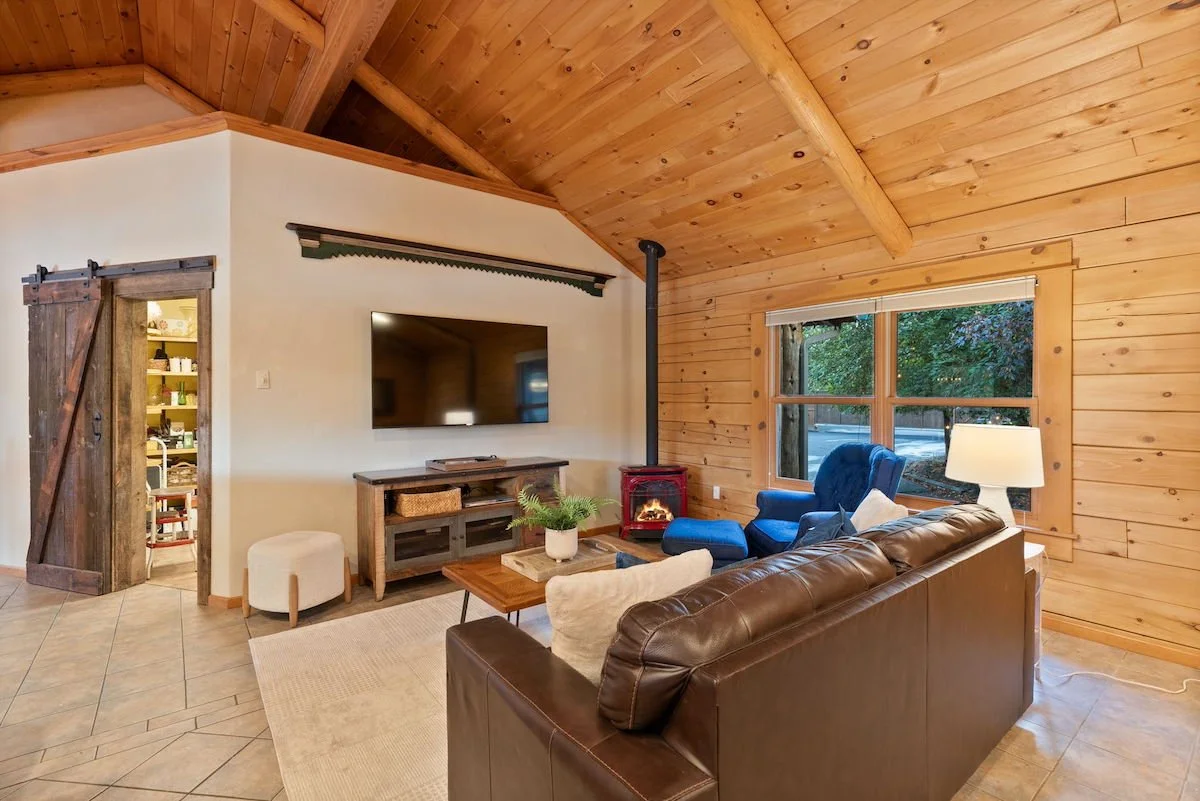 Living room with wood-paneled walls and ceiling, leather sofa, blue armchair, television, small fireplace, window showing greenery outside, and doorway revealing pantry.